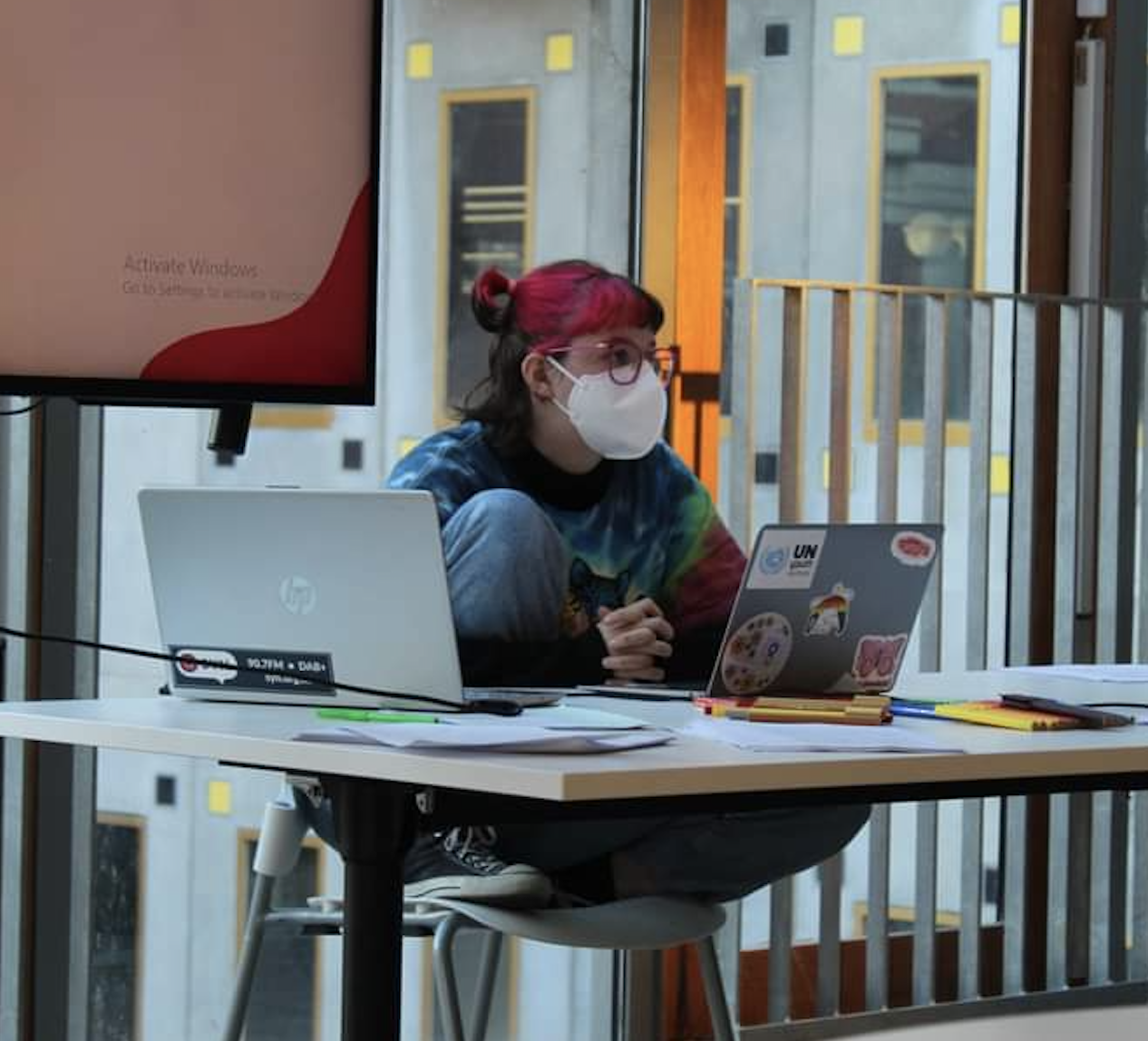 Photo of Is, a white trans person with pink and brown hair sitting behind a laptop at a computer.