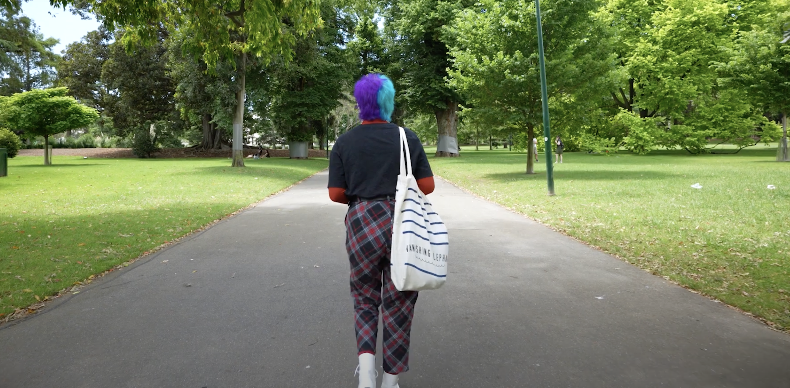 Photo of Is' back walking on a grey path in a green park with trees and grass. Is has blue and purple hair, carries a tote bag and wears black and red clothes