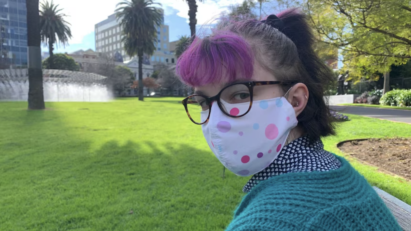Photo of Is, a white trans person with pink and black hair, brown glasses, a colourful fabric mask and blue clothes. They sit in a park with green grass, a water fountain, trees and buildings in their background and look at the camera.