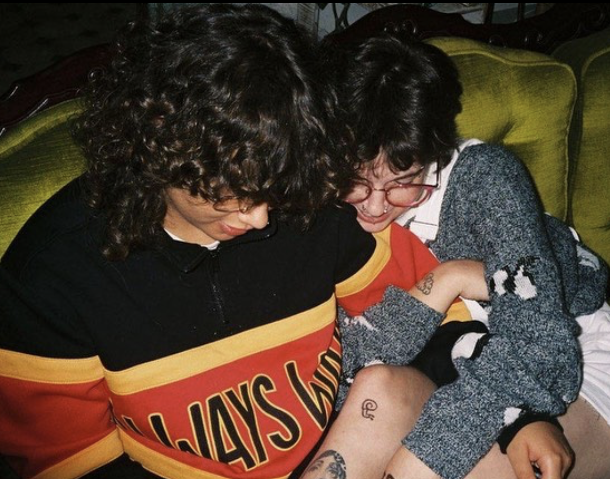 Photo of Is & Stevie cuddled up looking down. Stevie is a Brazilian man with curly brown hair and wearing an 